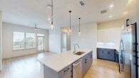 Located at 1345 14th St in Plano, TX, this thoughtfully designed two-bedroom, two-bathroom residence offers a contemporary open-concept layout across a single floor. The combined kitchen and living area serves as the heart of the home, featuring light hardwood-style flooring, recessed lighting, and a ceiling fan. The modern kitchen is equipped with stainless steel appliances including a French door refrigerator, electric range, and built-in microwave. Two-toned cabinetry — white upper cabinets contrasted with dark lower units — complements the clean white quartz countertops and a large kitchen island with pendant lighting. The living area opens to an outdoor balcony through a glass-panel door flanked by wide windows, flooding the space with natural light and offering pleasant outdoor views. Both bedrooms feature plush carpeting and windows with exterior views, providing a comfortable and quiet retreat. Each bedroom is paired with its own dedicated closet. The primary bathroom impresses with marble-look tile flooring, dual vanity sinks set in blue-grey cabinetry with black hardware, large mirrors, and a glass-enclosed walk-in shower with marble tile surround. In-unit laundry is also accessible from the main living corridor, adding everyday convenience to this well-appointed Plano residence.