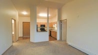 This single-floor residential space offers a thoughtfully laid-out interior that balances functionality with a warm, comfortable ambiance. The living area is finished with plush wall-to-wall carpeting that lends a cozy feel underfoot, while vaulted ceilings add a sense of openness and airiness throughout the space.

The kitchen is partially open to the main living area, separated by a half-wall with structural columns that maintain a visual connection between the two spaces. It features warm honey-toned wood cabinetry in abundance, providing ample storage. Black appliances, including a range, microwave, and dishwasher, offer a clean contrast against the light countertops and cabinetry. The kitchen floor transitions to a hardwood-style surface, clearly delineating the cooking zone from the carpeted living space.

A hallway visible from the main living area leads to additional rooms, with doors suggesting access to a bathroom and at least one additional room or closet. A front entry door with a warm wood finish is visible from the living area, contributing to the unit's welcoming character. Baseboard heating units are present along the walls, providing efficient climate control. Overall, the space maintains a neutral color palette of warm beiges and whites, creating a bright, clean, and inviting environment.