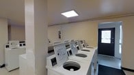 This single-floor laundry facility is designed for shared use, offering a straightforward and functional layout. A row of top-loading washing machines lines one wall, each unit clearly labeled with operational instructions for user convenience. The machines are arranged in a tidy sequence, providing ample capacity for multiple users. The room features neutral-toned walls and is illuminated by a flush-mounted overhead panel light that ensures bright, even lighting throughout the space. A fire extinguisher is mounted on the wall, reflecting attention to safety standards. A folding or utility surface appears along the back wall, offering a practical area for sorting or staging laundry. The entrance features a dark-framed door that allows natural light to filter in from outside, adding a secondary light source to the room. The overall ambiance is clean, utilitarian, and well-maintained, making it a practical shared amenity. The space is efficiently organized to accommodate laundry needs with minimal clutter.