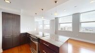 Located at 909 Walnut St in the heart of Kansas City, MO, this two-bedroom, two-bathroom residence offers a well-designed, contemporary floor plan with generous living space spread across a single level. The open-concept kitchen is a standout feature, showcasing rich dark-wood cabinetry, light granite countertops with a central island, stainless steel appliances including a French door refrigerator and slide-in range, and pendant lighting overhead. Warm hardwood flooring runs throughout the main living areas, complementing the clean, modern aesthetic. Large windows frame elevated views of the Kansas City skyline, flooding the living spaces with natural light. The two living areas provide flexible space for dining and lounging. The bedrooms are carpeted for comfort, with the spacious primary bedroom offering a well-sized walk-in closet with built-in shelving. The bathrooms are finished with granite-topped dark-wood vanities, tiled surrounds, and soaking tubs. Two dedicated closets provide ample storage throughout. The overall ambiance is sleek and urban, with a neutral color palette and high-quality finishes that reflect a modern, city-living aesthetic.