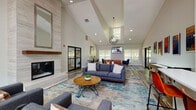 The clubhouse at Hunter Ridge in Peachtree Corners, Georgia, is a thoughtfully designed single-floor community gathering space that blends modern aesthetics with functional amenities. Soaring vaulted ceilings define the open-concept interior, creating a sense of grandeur throughout. Rich wood-look flooring, neutral gray walls, and vibrant area rugs anchor the various seating zones while maintaining a cohesive, contemporary feel.

The main lounge area serves as the heart of the clubhouse, anchored by a dramatic floor-to-ceiling tile fireplace with a wood mantel and a large mirror above — an unmistakable focal point. Comfortable sofas and accent chairs are arranged to encourage conversation, complemented by an overhead orb-style chandelier and recessed lighting. A wall-mounted flat-screen TV adds an entertainment dimension to this relaxed gathering zone.

Adjacent to the lounge is a casual dining nook with a wooden table and modern blue chairs, bathed in natural light from large windows that offer views of the outdoor pool and patio area. A bar-height counter with seating provides an additional space for informal gatherings near a kitchenette area.

The business center features a long wall-mounted desk with multiple computer workstations, stylish seating, and its own mounted television — ideal for residents seeking a productive workspace. A shuffleboard table rounds out the recreational offerings, adding a playful touch to this well-appointed community clubhouse.