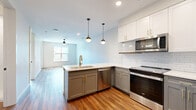 This thoughtfully designed one-bedroom, one-bathroom unit offers a well-organized single-floor layout with modern finishes throughout. Rich hardwood-style flooring flows seamlessly across the entire space, creating a warm and cohesive aesthetic.

The open-concept kitchen and living area serves as the heart of the unit. The kitchen is outfitted with two-tone cabinetry — white upper cabinets with gold hardware and gray lower cabinets — complemented by a classic white subway tile backsplash, quartz countertops, and a full suite of stainless steel appliances. A peninsula with a single-basin sink and stylish globe pendant lights overhead adds both function and visual appeal, while recessed lighting brightens the adjoining living area.

The bedroom is a comfortable, well-lit retreat featuring a large window with views of the surrounding building exterior and a ceiling fan for year-round comfort. A glass-panel door leads directly to a private balcony, allowing natural light to fill the space.

The walk-in closet provides ample storage with built-in shelving and hanging rods. The bathroom is a standout feature, boasting a bold navy-blue vanity with gold fixtures, a large mirror with lighted sconces, and a walk-in glass-enclosed shower with white tile surround and gold hardware accents.