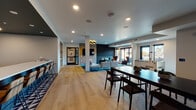This thoughtfully designed single-floor space offers a diverse range of amenity areas united by a cohesive contemporary aesthetic. The open-plan social lounge anchors the interior, featuring warm light wood flooring, recessed lighting, and a comfortable seating arrangement centered around a wall-mounted television and dark accent wall. Large windows flood the space with natural light and provide outdoor views, while a foosball table adds a recreational element. A long communal dining table offers additional gathering space nearby.

The co-working zone features a round conference table surrounded by tan leather task chairs, set atop a rich teal-toned patterned carpet. Adjacent private offices with glass-panel doors provide focused work options, and a built-in wooden bookshelf adds warmth and functionality. A sleek kitchenette serves the space, boasting light wood cabinetry, white countertops, open metal shelving, a tiled backsplash, and a bold geometric-patterned floor.

The fitness area is well-equipped with free weights, kettlebells, medicine balls, and a weight bench, set against a patterned accent wall. A seating nook showcases a striking panoramic cityscape photograph. The accessible restroom features dark mosaic tile walls with an artistic upper border. An outdoor courtyard with metal railings and gravel landscaping completes the offering.