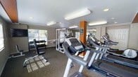 This fitness center is thoughtfully designed to support a full range of workout routines within a single, open-plan floor. The space is anchored by a row of ProForm treadmills positioned prominently in the center, offering residents easy access to cardiovascular training. A multi-station cable and weight machine occupies the mid-section of the room, providing versatile strength training options, while a dedicated free weights area along one wall features a well-stocked dumbbell rack paired with a workout bench, catering to those who prefer traditional strength exercises.

Natural light filters in through large windows along the exterior wall, creating a bright and energizing atmosphere. Full-length mirrors line one wall, visually expanding the space and allowing users to monitor their form during workouts. Overhead, a combination of elongated flush-mount light fixtures and recessed lighting ensures consistent, even illumination throughout. The flooring consists of durable, low-pile carpet suited for a fitness environment. An orange accent column adds a subtle pop of color, contributing to a modern and motivating aesthetic. The overall ambiance is clean, functional, and well-maintained.