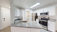 This well-appointed single-floor residence offers a clean, modern aesthetic defined by a consistent palette of crisp white walls, light wood-look flooring, and contemporary finishes throughout. The open-concept kitchen serves as a central highlight, featuring white shaker-style cabinetry, quartz countertops, and a peninsula with a built-in sink that opens toward the main living area. Stainless steel appliances — including a refrigerator, electric range, and over-the-range microwave — complement the bright, functional workspace, while flush-mounted ceiling lighting keeps the area well illuminated. Adjacent to the kitchen, a dedicated laundry room provides added convenience with side-by-side washer and dryer units and overhead shelving for storage. The living area is spacious and airy, benefiting from large sliding glass doors fitted with vertical blinds that invite natural light and provide access to the outdoors. The bedroom features dual windows with horizontal blinds, allowing for generous natural light and a comfortable, tranquil environment. The bathroom is well-equipped with a quartz-topped vanity, a large wall mirror, globe lighting, and a full tub-shower combination. A programmable thermostat and ample wall outlets are noted throughout, underscoring the unit's practical, move-in-ready design.