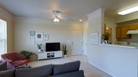 This single-floor residence offers a clean, contemporary layout designed for comfortable everyday living. The living room serves as the heart of the home, featuring plush carpeted flooring, crown molding, and a ceiling fan with integrated lighting that keeps the space well-lit and comfortable. A low-profile white media console anchors the entertainment area, complemented by an assortment of framed wall art and lush indoor plants that add a touch of warmth and personality to the space. A grey sofa provides a cozy focal point for the room. The entryway is defined by a classic paneled front door, tile flooring, and a half-wall partition that subtly separates the foyer from the adjoining areas, maintaining an open and airy feel throughout. Recessed lighting supplements the natural light filtering into the space, keeping the interior bright and inviting. The half-wall opens to what appears to be a kitchen or dining area, suggesting a connected, open-concept floor plan ideal for both relaxed living and entertaining. Neutral wall tones and consistent trim details tie the design together, creating a cohesive and welcoming atmosphere throughout this thoughtfully arranged single-level home.