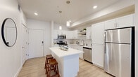 This thoughtfully designed single-floor residence offers 1 bedroom and 1 bathroom across a compact yet efficiently utilized floor plan. The open-concept kitchen is a standout feature, showcasing white shaker cabinetry, open display shelving, stainless steel appliances including a gas range, microwave, dishwasher, and refrigerator, and a central island with a white quartz countertop and bar seating. Pendant lighting adds a modern touch overhead, while light wood-tone flooring runs throughout the main living areas. The kitchen flows seamlessly into the living room, which is bathed in natural light from large windows and a sliding glass door. The bedroom offers plush carpet flooring, a sizable window for natural light, and ample space for a full furniture arrangement. The full bathroom features a tiled tub-shower combination, light wood-tone flooring, and a white vanity. A dedicated laundry room includes a full-size washer and dryer, wire shelving for storage, and additional built-in drawers — a practical and convenient feature. The hallway connects all areas of the home efficiently. Clean white walls and recessed lighting throughout lend the space a bright, modern, and cohesive aesthetic.