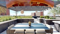 This rooftop terrace is a thoughtfully designed outdoor space that combines functional amenities with a refined aesthetic, offering an elevated urban retreat. The terrace is anchored by a striking curved pergola with a warm wood-paneled ceiling, beneath which string lights and a woven pendant fixture create an inviting ambiance for outdoor dining. A sleek dining table with upholstered slatted chairs sits at the heart of this sheltered area, framed by low concrete-clad seating walls that define the space.

Adjacent to the dining zone, a fully equipped outdoor kitchen features a stone-clad base housing a professional-grade gas grill, a pizza oven, and prep surfaces, all positioned beneath a modern open pergola with retractable elements. This cooking area is well-suited for hosting gatherings of various scales.

Raised planting beds filled with lush grasses, flowering plants, and low shrubs border the terrace perimeter, providing both greenery and a sense of enclosure. Large-format stone pavers in a neutral tone cover the floor throughout, contributing to the terrace's clean, contemporary character. Panoramic views of the surrounding urban skyline, neighboring buildings, and open sky complete the experience, with abundant natural light enhancing the open-air atmosphere year-round.