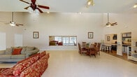 Located at 2335 Atkinson Rd in Biloxi, Mississippi, this single-floor space offers a well-organized layout comprising a living room and kitchen. The living room is a standout feature, characterized by dramatically vaulted ceilings that create an expansive, open atmosphere. Two ceiling fans provide comfortable airflow throughout the room, while recessed lighting adds warmth. Large windows and a glass door along one wall flood the space with abundant natural light and provide views of the surrounding greenery outdoors. Light-toned tile flooring extends throughout the living area, enhancing the bright and airy ambiance. A full-length wall mirror with a wood frame further amplifies the sense of depth and openness. The living room also accommodates a dining area, visible from the open layout, complete with a wooden table and chairs. The kitchen is efficiently designed, featuring dark navy cabinetry paired with granite-style countertops, a double stainless steel sink, and black appliances including a refrigerator and microwave. Wood-look flooring in the kitchen adds a warm contrast to the cabinetry. Upper and lower cabinets provide ample storage. Together, the rooms offer a cohesive, functional space with a clean, welcoming aesthetic suited for a variety of uses.