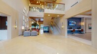 Located at 3991 NW 82nd Dr in Cooper City, Florida, this upscale residential amenity facility showcases a sophisticated, contemporary design throughout its expansive single-floor layout. The grand entrance lobby immediately impresses with soaring double-height ceilings, warm wood-paneled accents, large-format porcelain tile flooring, and a striking open staircase with glass railings leading to an upper mezzanine level. Pendant lighting installations and a dramatic water feature serve as centerpiece focal points, while lush indoor trees add a natural, resort-like ambiance. A generously sized lounge area features curated seating arrangements with navy blue and neutral-toned furnishings, layered rugs, and gallery-style wall art. An adjacent social lounge and café-style seating area offers additional gathering space with coffered ceilings, recessed lighting, and views to the outdoors. The dedicated conference room is appointed with a solid wood communal table, gold-toned geometric chandelier, built-in cabinetry, and a mounted display screen, with direct access to an outdoor terrace. A business center nook provides workstation access, while a well-organized mail room features abundant individual mailboxes accented with tropical greenery. Throughout, the design balances warmth and modernity with consistent use of wood, glass, and neutral tones.