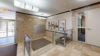 Located at 28 Elizabeth St N in Mississauga, Ontario, this building lobby serves as a welcoming and functional common entrance for residents and visitors alike. The space is defined by its distinctive stone-clad walls in warm beige and tan tones, lending a classic yet refined character throughout. Travertine-style tile flooring extends across the entire area, complemented by textured entry mats that provide practical transitions between zones.

A notable architectural feature is the split-level layout, with a short staircase — equipped with sleek stainless steel and glass railings — leading up to the elevator vestibule. The elevator itself is centrally positioned and easily accessible, with an additional door nearby offering further building access. A bank of individual resident mailboxes is mounted along one wall, serving as a key functional component of the lobby.

The space is illuminated by a combination of flush-mounted ceiling fixtures and pendant-style hanging lights, creating a bright and inviting ambiance. A textured popcorn ceiling with crown molding detailing adds a traditional finishing touch. Three vibrant, colorful framed artworks are displayed prominently on the wall, introducing a lively and modern aesthetic contrast to the stone surroundings. A glass-paneled entrance vestibule at the front allows natural light to filter in, enhancing the overall brightness of this well-maintained and thoughtfully designed lobby.