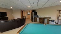 This single-level recreational space is thoughtfully designed for entertainment and leisure. At its center sits a full-size pool table with a teal felt surface, serving as the focal point of the room and providing ample space for gameplay. Adjacent to the table is a comfortable lounge area furnished with a plush leather sectional sofa, creating a relaxed setting for guests to unwind between games.

The kitchenette and wet bar area is well-equipped, featuring a stainless steel refrigerator, microwave, and a sink with countertop space finished in a dark granite-style surface — offering convenient access to refreshments without leaving the room. Cabinetry below the counter provides additional storage.

The space is finished with neutral-toned carpeting and warm beige walls, contributing to a cozy yet functional atmosphere. A drop ceiling with recessed and panel lighting ensures consistent, even illumination throughout. Interior doors lead to adjoining areas, adding to the overall connectivity of the layout. This space is well-suited for hosting gatherings, game nights, or casual socializing, combining practical amenities with a welcoming recreational atmosphere.