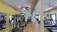 This fitness center offers a thoughtfully designed workout environment suited for a wide range of exercise routines. The space features warm hardwood-style flooring throughout, providing a clean and durable surface ideal for both machine-based and open-floor training. Bright recessed lighting is complemented by a ceiling fan, ensuring the area stays well-lit and comfortable during workouts.

The strength training section is well-appointed with a multi-station cable and weight machine, an adjustable workout bench, and a fully stocked dumbbell rack offering a range of free weights. Wall-mounted mirrors line portions of the space, allowing users to monitor their form during exercises. Suspension training straps are also available, adding versatility to bodyweight workouts.

The cardio area features treadmills and additional equipment positioned toward the perimeter, making efficient use of the floor plan. The yellow accent walls add an energetic, motivating atmosphere to the interior. Structural columns define zones within the open layout without interrupting the flow of the space. A bulletin board displays community notices, adding a welcoming, community-oriented touch. Natural light filters in from an adjacent area visible through a doorway, further brightening the overall environment.