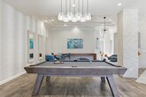 Located at 7450 Northrop Dr in Riverside, California, this single-story community clubhouse offers a thoughtfully designed gathering space that blends modern aesthetics with functional amenities. The space is anchored by an open-plan lounge area with soaring tray ceilings, recessed lighting, and statement sputnik chandeliers that add a contemporary flair. Wide-plank wood-look flooring flows throughout, unifying the space with a warm, cohesive feel. A striking stacked-stone pillar serves as a central architectural feature, framing a cozy seating area complete with a fireplace and wall-mounted television. Large French doors and expansive windows flood the lounge with natural light and offer views of the outdoor pool area. The adjacent game room is well-appointed with a billiard table, shuffleboard, high-top seating, and additional lounge chairs, making it ideal for social gatherings. Subtle striped wallcovering adds texture and depth to this area. The fully equipped kitchen features warm wood cabinetry, stainless steel appliances including a gas range and refrigerator, a bold chevron-patterned tile backsplash, white quartz countertops, and a peninsula with bar stool seating beneath globe pendant lights — a functional and stylish space for entertaining.