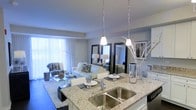 This single-floor residence showcases a thoughtfully designed open-concept layout that seamlessly connects the kitchen and living areas. The kitchen is anchored by a generous island topped with light granite countertops, a stainless steel undermount sink, and a contemporary faucet. Two sleek pendant lights hang above the island, providing focused task lighting while contributing to the modern aesthetic. White cabinetry and quality appliances complement the clean, polished look of the space.

The living area flows naturally from the kitchen, creating an inviting and cohesive environment. Large floor-to-ceiling windows dressed with sheer white vertical blinds allow an abundance of natural light to fill the room, enhancing its airy atmosphere. The living space is generous enough to accommodate a full seating arrangement, with room for accent chairs and a coffee table. Decorative full-length mirrors along one wall add depth and elegance. Framed wall art, decorative vases, and coordinated furnishings complete the refined, contemporary ambiance throughout this well-appointed residence.