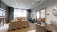 This refined single-floor space blends professional functionality with a warm, inviting atmosphere. The main lounge area is thoughtfully appointed with a plush beige sofa accented by colorful throw pillows, complemented by a pair of teal upholstered armchairs arranged atop a patterned area rug. A decorative starburst mirror serves as a focal point on the neutral gray wall, adding an elegant design touch. Large windows dressed with layered roller shades and floor-length gray drapes allow for abundant natural light while maintaining privacy. Recessed ceiling lighting provides consistent, well-distributed illumination throughout the space. Adjacent to the lounge, a glass-paneled conference room is visible, outfitted with a long meeting table and ergonomic leather chairs, making it well-suited for professional gatherings. A curved reception or service counter anchors the transitional area between the lounge and the conference room, contributing to a seamless flow across the space. The overall palette of soft grays, warm beiges, and cool teal accents creates a calm yet professional ambiance, ideal for a client-facing business environment such as a medical office, financial services firm, or corporate suite.