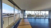 This single-floor residence showcases a thoughtfully designed interior centered around an abundance of natural light and panoramic urban views. A clean, white-walled hallway with dark hardwood flooring serves as the entry corridor, lined with doors on either side and a ceiling-mounted light fixture overhead. The hallway leads directly toward the main living area, where the design opens dramatically.

The main living space is defined by an expansive wall of floor-to-ceiling glass panels and sliding doors that wrap around the room, offering unobstructed views of the city skyline and what appears to be a body of water beyond. Sunlight streams through the glazing, casting strong natural light across the warm-toned hardwood floors. Sheer panel curtains are installed along the glass wall, offering a degree of privacy and light control while maintaining the open, airy atmosphere. A balcony or terrace is accessible through the sliding glass doors, further connecting the interior to the surrounding urban landscape. The overall aesthetic is modern and minimal, emphasizing clean lines, neutral tones, and the architectural feature of its remarkable view-oriented design.