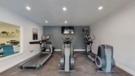 Located at 2255 Country Corners Dr in Columbus, Ohio, this fitness center offers a well-appointed, single-floor exercise space designed to support an active lifestyle. The room is finished with a textured ceiling and recessed lighting that evenly illuminates the area, creating a bright and functional environment for workouts. A bold slate-blue accent wall serves as the focal point of the space, anchored by a wall-mounted flat-screen television — ideal for staying entertained during cardio sessions. The flooring consists of durable, patterned carpet tiles well-suited for a high-traffic fitness environment. The facility is equipped with a selection of cardio machines, including a treadmill, a stair climber, and elliptical trainers, providing a variety of options for aerobic exercise. A stability ball is also available, adding flexibility for supplementary strength and balance training. Motivational wall art completes the aesthetic, contributing to an energizing atmosphere. The layout is thoughtfully organized to allow comfortable movement between equipment. Clean white walls contrast with the accent wall to keep the space feeling open and inviting. Overall, this fitness center delivers a practical and well-maintained amenity for residents at this Columbus location.