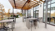 This contemporary single-floor space blends modern architecture with functional design, offering a seamless transition between indoor and outdoor environments. The standout feature is a spacious covered outdoor patio, sheltered by a striking wood-beam pergola with decorative string lighting overhead. The patio is furnished with sleek concrete high-top tables and metal bar stools, creating an inviting setting for gathering or dining. Large floor-to-ceiling glass windows and black-framed doors line the exterior wall, flooding the interior with natural light while maintaining a strong visual connection to the outdoors. Views from the patio extend to a well-landscaped surroundings with trees and open streetscapes beyond.

Inside, a clean, minimalist aesthetic prevails throughout. White large-format tile flooring runs continuously through the corridors, which are lined with neutral-toned walls and subtle artwork accents. Recessed lighting provides even illumination throughout the hallways, which lead to additional interior rooms accessed through warm wood-toned doors. The entry vestibule features a glass door with transom window, reinforcing the building's commitment to natural light and transparency. Overall, the space presents a polished, professional atmosphere suited for commercial or mixed-use occupancy.