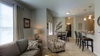 This single-floor residence offers a thoughtfully designed open-concept layout that seamlessly connects the living and dining areas, creating a comfortable and inviting atmosphere. The living room features vaulted ceilings that lend a sense of spaciousness, complemented by soft, neutral wall tones and plush carpeted flooring. A coordinated seating arrangement, including a patterned armchair and sofa with matching accent pillows, anchors the space alongside a decorative table lamp and framed wall art, adding warmth and character. The dining area flows naturally from the living room and is furnished with a round table and upholstered chairs, accented by an eye-catching metallic wall decoration. Natural light filters through window treatments, brightening the space. A hallway visible from the main living area leads toward additional rooms, with glimpses of a well-lit vanity area suggesting a private bathroom with dedicated lighting. The open sightlines throughout the floor plan enhance the overall sense of flow and connectivity, making this a practical yet aesthetically refined space suitable for everyday living and entertaining alike.
