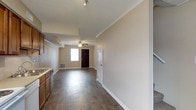 This two-floor residence offers a thoughtfully arranged layout that balances functionality and comfort. The main level features rich, dark hardwood flooring that runs throughout, providing a warm and cohesive foundation for the open living space. Crown molding lines the ceilings, adding a refined architectural detail that elevates the overall aesthetic.

The living and entry area is anchored by a ceiling fan and benefits from natural light streaming through a front-facing window. A dark wood entry door creates a welcoming focal point at the entrance. Track lighting near the kitchen zone provides additional illumination, complementing the warm-toned cabinetry visible along the kitchen perimeter.

The open-concept flow between the kitchen and living areas makes the main floor feel spacious and connected, ideal for everyday living. Neutral wall tones throughout create a clean, versatile backdrop that adapts easily to a variety of interior styles.

With two floors of living space, residents have ample room for dedicated sleeping, living, and utility areas. The combination of classic finishes — crown molding, hardwood floors, and traditional cabinetry — gives the home a timeless character, while practical features like built-in ventilation registers ensure year-round comfort throughout the space.