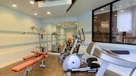 This fitness center is designed to support a comprehensive workout experience within a compact, efficiently organized space. The room features light blue walls accented with subtle horizontal stripes, polished concrete flooring, and recessed ceiling lights that create a bright and energizing atmosphere. A large floor-to-ceiling mirror spans one wall, visually expanding the space and allowing users to monitor their form during exercise. The equipment selection is well-rounded, including an elliptical machine for cardiovascular training, a multi-station cable weight machine for strength training, an adjustable workout bench with free weights, and additional resistance-based equipment. A large window on one side of the room connects the fitness area to an adjacent space featuring exposed brick walls, adding a warm, urban aesthetic contrast to the clean, modern interior. The ceiling incorporates a skylight or upper-level opening that introduces additional natural light into the room. Overall, the fitness center is thoughtfully arranged to maximize functionality, offering residents or users access to both cardio and strength training options within a clean, well-lit, and motivating environment.