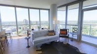 This sophisticated high-rise residence offers an impressive living experience defined by wraparound floor-to-ceiling glass windows that flood the interior with natural light and frame sweeping panoramic views of the city skyline and lush tree canopy below. The open-plan living area is designed with a modern sensibility, anchored by rich hardwood flooring and a neutral color palette that allows the stunning views to take center stage. A sleek, tufted white sofa paired with a leather accent chair creates an inviting seating arrangement, complemented by a cowhide area rug that adds texture and warmth to the space. Thoughtfully placed accent tables and a tall floor lamp provide both function and style. The corner positioning of the living space maximizes the expansive outlook, offering dynamic perspectives in multiple directions. The overall ambiance is bright, airy, and contemporary, with clean architectural lines punctuated by curved structural columns that add a distinctive design element. The seamless connection between the interior and the vast outdoor views beyond the glass creates a sense of openness and elevation, making this a truly elevated urban living environment.