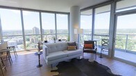 This sophisticated high-rise residence offers an impressive living experience defined by wraparound floor-to-ceiling glass windows that flood the interior with natural light and frame sweeping panoramic views of the city skyline and lush tree canopy below. The open-plan living area is designed with a modern sensibility, anchored by rich hardwood flooring and a neutral color palette that allows the stunning views to take center stage. A sleek, tufted white sofa paired with a leather accent chair creates an inviting seating arrangement, complemented by a cowhide area rug that adds texture and warmth to the space. Thoughtfully placed accent tables and a tall floor lamp provide both function and style. The corner positioning of the living space maximizes the expansive outlook, offering dynamic perspectives in multiple directions. The overall ambiance is bright, airy, and contemporary, with clean architectural lines punctuated by curved structural columns that add a distinctive design element. The seamless connection between the interior and the vast outdoor views beyond the glass creates a sense of openness and elevation, making this a truly elevated urban living environment.