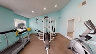 This single-floor amenity space offers residents a comprehensive set of recreational facilities designed to support an active and comfortable lifestyle. The fitness center is bright and functional, featuring light aqua-painted walls, carpeted flooring, and recessed lighting throughout. It is equipped with a multi-station cable weight machine, a treadmill, and additional cardio equipment, complemented by full-length mirrors along one wall that enhance the sense of space. Natural light filters in through slatted windows, and a built-in bench with accent pillows provides a convenient resting area.

Outdoors, the community shines with a generously sized, free-form swimming pool accompanied by an adjacent circular spa, both finished in striking turquoise tile. Lounge chairs are positioned poolside, and the area is beautifully framed by tall, mature palm trees that lend a resort-like ambiance under clear blue skies. The surrounding architecture features a warm stucco exterior consistent with a Southwestern aesthetic.

A dedicated children's playground rounds out the amenities, featuring a multi-slide play structure with green accents set on a sandy surface, offering a safe and engaging outdoor space for younger residents.