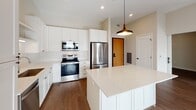 Located at 1416 Second St SW in Rochester, Minnesota, this modern single-floor unit offers a thoughtfully designed open-concept layout. The kitchen is a standout feature, equipped with stainless steel appliances including a refrigerator, range, and built-in microwave, complemented by crisp white shaker cabinetry, a subway tile backsplash, and a generous island topped with quartz countertops. A stylish pendant light anchors the kitchen space, which flows seamlessly into the bright living area. The living room is bathed in natural light through large black-framed windows and a glass door that opens to a balcony, offering views of the surrounding Rochester cityscape. Exposed concrete columns add an industrial architectural accent throughout the common areas. Hardwood-style flooring runs through the main living spaces, while carpeting provides warmth in the bedrooms and walk-in closet, which features wire shelving for ample storage. The primary bathroom is well-appointed with a dual-sink vanity, white quartz countertops, white cabinetry with matte black hardware, and a large wall mirror. A secondary bathroom provides additional functionality with similar finishes. Recessed lighting throughout ensures a clean, well-lit interior, while the neutral warm-toned walls create a welcoming and versatile ambiance.