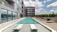 This thoughtfully designed outdoor amenity level in Nashville, TN (37209) offers a compelling array of communal features set against the backdrop of a modern multi-story building clad in dark gray brick and metal panel siding with large-format windows and private balconies above.

At the heart of the space is a generously sized rectangular swimming pool with crystal-clear blue water, flanked by rows of lounge chairs and chaise seating ideal for sunbathing. Adjacent to the pool, a seating area with navy blue cushioned chairs surrounds a fire pit table, creating an inviting gathering spot for cooler evenings. Large floor-to-ceiling sliding glass doors connect the pool deck to an interior space, allowing for seamless indoor-outdoor flow.

The courtyard transitions into a lush artificial turf lawn, where string lights are strung overhead between posts, lending a warm, festive ambiance day and night. A built-in outdoor kitchen with a grill and brick surround anchors one end of the lawn, accompanied by a dining table and a sculptural hanging egg chair. Vibrant geometric murals with bold splashes of yellow, blue, and orange animate the perimeter walls, adding an artistic, urban energy to the setting. Mature evergreen trees line the boundaries, providing both privacy and a touch of greenery throughout this well-appointed outdoor retreat.