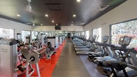 This expansive, single-floor fitness center is thoughtfully designed to accommodate a full range of workout needs. The space is divided into distinct zones, each with its own flooring that visually separates the areas — bold red and orange rubber flooring marks the strength and free weights sections, while grey tile defines the cardio zone. A long row of treadmills, ellipticals, and stair climbers lines one wall, all positioned to face large picture windows that let in generous natural light and offer views of lush outdoor greenery and the surrounding property. The strength training area is outfitted with a comprehensive selection of cable machines, weight benches, and plate-loaded equipment from brands including Cybex and True. A dedicated free weights section features a full rack of dumbbells and stability balls, complemented by floor-to-ceiling mirrors that enhance the sense of space throughout. A built-in service station with dark wood cabinetry, open shelving stocked with fresh towels, a sink, and a mini-refrigerator adds a convenient, upscale touch. Ceiling fans and recessed lighting are distributed throughout, while mounted televisions provide entertainment. The dark ceiling provides a striking contrast to the white walls, giving the space a modern, polished aesthetic.