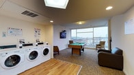 This single-floor amenity space combines laundry facilities with a recreation area, offering residents a functional and social environment. The laundry section features multiple front-loading washers and dryers arranged along one wall, accompanied by posted usage instructions and hardwood flooring underfoot. A skylight overhead floods this area with natural light, enhancing the bright, open atmosphere.

Transitioning into the recreational zone, the flooring shifts to patterned carpet tiles, delineating the two distinct areas. A full-sized pool table serves as the centerpiece of the recreation space, positioned to take full advantage of the room's generous proportions. A wall-mounted flat-screen television is installed nearby, adding to the entertainment options available.

The highlight of the recreation area is a expansive floor-to-ceiling window that spans nearly the full width of the exterior wall, framing sweeping views of the surrounding cityscape and rooftops beyond. A pair of comfortable armchairs are positioned near the window, creating an inviting seating nook. Recessed lighting and a ceiling-mounted fixture provide additional illumination throughout the space, ensuring functionality during all hours.