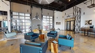 This single-floor clubhouse makes an immediate impression with its industrial-modern design language, blending soaring ceilings, exposed black ductwork, and rich polished concrete floors that reflect the abundant natural light pouring in through oversized glass garage doors and floor-to-ceiling windows. An aviation theme runs throughout, highlighted by a striking propeller chandelier, vintage aircraft artwork, and curated décor that lends the space a distinctive, memorable character.

The main lounge is generously proportioned, furnished with teal and warm-toned leather seating arranged around natural wood accent tables, creating inviting conversation areas beneath dramatic pendant lighting. Large glass garage doors open the lounge to the outdoors, reinforcing a seamless indoor-outdoor connection.

A dedicated game room features a rustic wood pool table, bar-height seating, and expansive glazed walls that frame palm-lined outdoor views, flooding the area with daylight. Adjacent to this is a stylish coffee and refreshment bar, complete with a solid wood communal table, leather counter stools, and a built-in service counter framed by a steel-paned interior window.

A reception area greets visitors with floor-to-ceiling glazing along one side, a staffed desk, and semi-private partitions with industrial steel framing. A media lounge nook offers wall-mounted entertainment and ambient lighting for a more relaxed, intimate setting. Together, these areas form a cohesive, amenity-rich environment designed for both socializing and productivity.