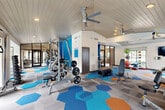 Located at 12951 Briar Forest Dr in Houston, TX, this fitness center offers a generously sized, single-floor workout facility designed to support a full range of exercise activities. The space is immediately striking thanks to its vaulted, tongue-and-groove white ceiling, which creates an open and airy atmosphere enhanced by dual ceiling fans that promote airflow throughout the room. A bold blue accent wall adds a vibrant, energetic character to the interior, complementing the eye-catching geometric hexagonal carpet tiles in shades of blue, orange, and gray that cover the entire floor. Large floor-to-ceiling glass windows and panel doors allow abundant natural light to flood the space while offering views of the surrounding outdoor area. The fitness center is comprehensively equipped with a dedicated cardio zone featuring treadmills and elliptical machines, as well as a strength training area outfitted with multi-station cable machines, free weights, a weight rack, and adjustable benches. Additional amenities include stability balls, resistance equipment, and mirrors along the walls to assist with form. Recessed lighting supplements the natural light, ensuring the space is well-lit at all times. The overall ambiance is modern, motivating, and functional, making it a well-rounded facility for residents seeking a convenient on-site fitness experience.