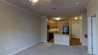This single-floor unit showcases an open-concept design that seamlessly connects the living area to the kitchen. The living space features neutral-toned carpet and warm beige walls accented by classic crown molding, creating a clean and welcoming atmosphere. A ceiling fan provides both practical ventilation and ambient lighting overhead, while a ceiling-mounted HVAC vent ensures year-round comfort.

The kitchen is positioned at the rear of the open layout and is equipped with warm-toned wood cabinetry offering generous storage. Black appliances, including a refrigerator, range, and microwave, provide a striking contrast against the lighter cabinetry. A peninsula with granite-style countertops extends into the space, creating a natural divide between the kitchen and living area while offering additional prep or dining space.

The kitchen floor transitions to hardwood-style flooring, distinguishing it from the carpeted living area. A utility sink is positioned along the kitchen wall beneath additional cabinetry. Near the entry, a white door with paneled detailing serves as the front entrance, completing the functional and straightforward layout of this well-organized single-floor space.