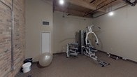 This fitness room combines an industrial aesthetic with practical workout functionality. The space is defined by its striking exposed brick wall along one side and an open ceiling structure featuring wooden beams and visible pipes and ductwork, lending the room a raw, warehouse-style character. Pendant lighting provides focused illumination throughout the area, complementing the neutral, carpeted flooring underfoot.

The room is equipped with multiple cardio machines, including treadmills and an elliptical trainer, positioned to allow comfortable use and movement between equipment. A multi-station strength training machine occupies one corner of the room, offering a variety of resistance exercises in a compact footprint. A stability ball and free weights round out the available equipment, making the space suitable for diverse workout routines.

A wall-mounted flat-screen television faces the cardio area, providing entertainment during workouts. The room is accessed through a solid wood door and features a ventilation grille for climate control. The neutral-toned walls contrast with the exposed brick, creating a balanced and functional atmosphere. Overall, this fitness room provides a well-equipped, utilitarian space with distinctive architectural character suited for daily exercise needs.