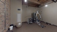 This fitness room combines an industrial aesthetic with practical workout functionality. The space is defined by its striking exposed brick wall along one side and an open ceiling structure featuring wooden beams and visible pipes and ductwork, lending the room a raw, warehouse-style character. Pendant lighting provides focused illumination throughout the area, complementing the neutral, carpeted flooring underfoot.

The room is equipped with multiple cardio machines, including treadmills and an elliptical trainer, positioned to allow comfortable use and movement between equipment. A multi-station strength training machine occupies one corner of the room, offering a variety of resistance exercises in a compact footprint. A stability ball and free weights round out the available equipment, making the space suitable for diverse workout routines.

A wall-mounted flat-screen television faces the cardio area, providing entertainment during workouts. The room is accessed through a solid wood door and features a ventilation grille for climate control. The neutral-toned walls contrast with the exposed brick, creating a balanced and functional atmosphere. Overall, this fitness room provides a well-equipped, utilitarian space with distinctive architectural character suited for daily exercise needs.