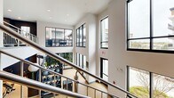 Located at 2702 Pelham Rd in Greenville, SC, this two-story fitness facility offers a comprehensive and well-appointed workout environment spread across two generously sized floors. The upper level is bathed in natural light from large floor-to-ceiling windows, offering views of the surrounding community and creating an open, airy atmosphere. This floor features treadmills, elliptical machines, and additional cardio equipment positioned to take advantage of the abundant daylight, alongside strength training stations and open workout areas. A staircase with sleek metal railings connects the two levels, with the double-height window wall creating a dramatic, light-filled transition between floors. The ground floor is equally well-equipped, housing cable machines, spin bikes, stability balls, resistance band racks, and a variety of free weights displayed on dedicated dumbbell racks. Accent walls in deep navy blue add a modern, energetic character throughout. A dedicated studio room features full-length mirror walls and light wood-toned flooring with linear ceiling lighting, ideal for stretching or group training. Support amenities include a water fountain and locker area. Ceiling fans are installed throughout for enhanced comfort, complemented by recessed lighting across both levels.