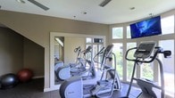 This fitness center is designed to provide a motivating and functional workout environment. The space is outfitted with a row of professional-grade Precor cardio machines, including ellipticals and a stair climber, arranged to take full advantage of the room's standout feature: a expansive floor-to-ceiling window wall that floods the space with natural light and offers refreshing views of surrounding greenery. The bright, open atmosphere created by the generous glazing makes the space feel airy and energizing during workouts. A large wall-mounted mirror on the opposite side further amplifies the sense of space while allowing users to monitor their form. Wall-mounted flat-screen televisions are positioned for easy viewing from the cardio equipment, enhancing the overall workout experience. The flooring features durable, exercise-appropriate tile, and the neutral warm-toned walls contribute to a clean and focused aesthetic. Recessed and flush-mount ceiling lighting supplements the abundant natural light, ensuring the space remains well-lit at all hours. The single-floor layout keeps everything accessible and straightforward, making this a practical and welcoming facility for fitness enthusiasts.