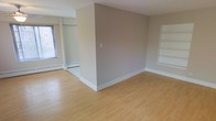 This single-floor space offers a clean, functional layout defined by light hardwood flooring that runs throughout, providing a warm and cohesive aesthetic. The open-concept design is partially divided by a central wall, which creates a natural separation between living areas while maintaining an airy, connected feel. Neutral tones across the walls complement the flooring and contribute to a bright, understated atmosphere.

One section of the space features a window that allows natural light to filter in, illuminating the tile flooring in that area and highlighting the baseboard heating unit positioned beneath it. A recessed closet or storage alcove with a track-style opening adds practical built-in storage to this zone.

On the opposite side, a set of built-in wall shelves provides additional storage or display space, adding architectural interest to an otherwise minimalist interior. The white baseboards and trim offer clean contrast against the neutral walls throughout.

Overall, the space presents a straightforward, well-maintained interior suited for versatile use, with practical features integrated into a simple, uncluttered design.