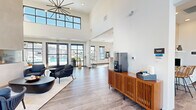 Located in Colorado Springs, CO, this single-floor community space offers a well-appointed, open-plan layout that seamlessly integrates a kitchen and living area. Dramatic soaring ceilings define the living area, amplifying the sense of openness and volume throughout the space. Large black-framed windows and glass double doors flood the interior with natural light while framing views of the surrounding Colorado landscape, including a pool area visible from the main gathering space.

The living area is anchored by a sleek corner fireplace that rises to the ceiling, providing a striking focal point. Multiple seating arrangements—including a tan leather sofa, woven lounge chairs, and accent tables—create versatile zones for gathering and relaxation. Durable wood-look flooring runs throughout, complementing the clean white walls adorned with contemporary artwork.

The kitchen features a granite-topped island with bar seating illuminated by industrial-style pendant lights. White cabinetry, glossy subway tile walls, open shelving, and stainless steel appliances—including a full-size refrigerator and microwave—complete the functional yet stylish culinary area. A coffee station is also present near the main lounge. The space presents a modern, welcoming atmosphere well-suited for community use.