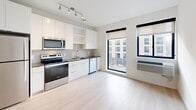 Located at 4046 E 46th St in Minneapolis, this single-floor unit presents a clean, modern aesthetic defined by light wood-style flooring, crisp white walls, and a cohesive contemporary design throughout. The open-concept kitchen is a central highlight, featuring white cabinetry, stainless steel appliances including a range, microwave, dishwasher, and refrigerator, along with a subway tile backsplash and track lighting overhead. Large black-framed windows flood the living area with natural light and offer views of the surrounding neighborhood, contributing to the bright and airy atmosphere. A wall-mounted HVAC unit provides climate control directly within the living space. The layout flows efficiently from the kitchen and living area through a connecting hallway, where a dark-paneled sliding door opens to a dedicated closet outfitted with wire shelving and hanging rods for organized storage. The bathroom maintains the unit's modern sensibility, featuring a light taupe vanity with a white countertop, a large frameless mirror, chrome fixtures, and a combined tub and shower. Recessed lighting is used consistently throughout, enhancing the clean, uncluttered feel of the space. The overall design prioritizes simplicity, functionality, and a well-lit environment.