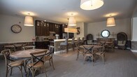 Located at 400 S Military Hwy in Virginia Beach, Virginia, this single-floor community space is thoughtfully designed to foster comfort and social connection. The open-concept layout flows seamlessly between distinct functional areas, all unified by a warm, neutral color palette accented with coastal-inspired teal and earthy tones throughout.

The central lounge area anchors the space with a generous sectional sofa in rich teal, complementary patterned accent chairs, and a stone fireplace column that adds architectural character. Recessed lighting illuminates the plush carpeted flooring, while framed coastal artwork enhances the relaxed ambiance.

Adjacent to the lounge, a dining area offers round tables with cross-back chairs beneath a pendant light fixture. Large windows dressed with teal valances and blinds flood the room with natural light and provide views of the surrounding exterior.

The fully equipped kitchen features dark espresso cabinetry extending to the ceiling with glass-front upper doors, a mosaic tile backsplash, light stone countertops, and stainless steel appliances including a range, microwave, and French door refrigerator. Light wood-tone flooring grounds the space with a clean, modern contrast.

A transitional corridor connects the areas, featuring cozy seating nooks with patterned accent chairs, decorative wall art, and access to additional areas. The overall design balances functionality with an inviting, well-appointed aesthetic.