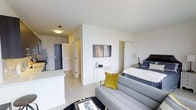 This well-appointed studio presents a thoughtfully designed open-plan layout that efficiently combines living, sleeping, and kitchen functions within a single, cohesive space. The interior is finished in a clean, contemporary palette of crisp white walls and smooth light-toned flooring, creating a bright and airy atmosphere throughout.

The living area features a comfortable grey sofa accented with a bold mustard yellow cushion, paired with a sleek coffee table — a stylish and functional arrangement that anchors the space. A wall-mounted flat-screen television serves as a focal point between the living and sleeping zones, which flow naturally into one another. The sleeping area is neatly defined by a well-sized bed dressed in neutral tones, complemented by a bedside lamp and side table.

The kitchen is positioned at the perimeter of the open space, featuring modern cabinetry and a clean countertop, offering practicality without compromising the open feel of the unit. A short hallway near the entry leads to the bathroom and provides useful storage access, while a front door with a secondary door visible nearby adds a sense of depth and separation from the corridor. Overall, the space offers a clean, modern, and functional living environment with a cohesive design aesthetic.