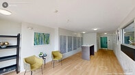 Located at 2504 NC-54 in Durham, NC, this single-floor lobby and hallway space serves as the welcoming entry point for the building. The interior is designed with a clean, contemporary aesthetic, featuring light hardwood flooring throughout that complements the bright white walls and ceilings, creating a fresh and airy atmosphere.

Upon entering through the floor-to-ceiling glass double doors, visitors are greeted by an open, well-lit reception area. A sleek reception desk with a dark wood base and white countertop provides a functional check-in point, while a large pass-through window connects the lobby to an adjacent office area, offering visibility into the back-of-house operations.

The hallway extends further into the building, where a comprehensive mailroom wall houses a large bank of individual resident mailboxes. A small seating area nearby, furnished with two upholstered chairs and a side table, offers a comfortable spot for residents. Colorful artwork adorns the walls, adding personality and vibrancy to the space.

A secondary secured entry door with keypad access provides controlled access further into the building. The space is efficiently lit with flush-mount ceiling fixtures and benefits from ample natural light streaming through the expansive glazed entrance, offering pleasant views of the landscaped parking area and surrounding greenery outside.