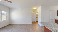 This single-story unit is thoughtfully designed with a clean, modern aesthetic throughout. The open-concept kitchen serves as a central feature, showcasing rich medium-toned wood cabinetry paired with light granite countertops, stainless steel appliances including a range, microwave, and refrigerator, and a kitchen island with a built-in sink — offering both functionality and a natural gathering point. Wood-look plank flooring runs through the main living and kitchen areas, lending a cohesive and easy-to-maintain finish.

The living area is generously sized, illuminated by natural light from windows with blinds and a glass exterior door, and fitted with a ceiling fan overhead. A secondary living or dining space connects seamlessly, also featuring wood-look flooring and a louvered interior door leading to additional rooms. The bedroom is carpeted and receives natural light through a window with blinds, and includes a ceiling fan. The bathroom features a full tub-shower combination, toilet, and wood-look tile flooring. A dedicated laundry area houses a full-size top-load washer and dryer with overhead shelving, completing the unit's practical amenities.