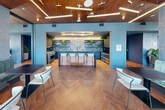 This thoughtfully designed amenity lounge occupies a single floor and offers a generous, open layout divided into distinct functional zones. The entry corridor features herringbone wood flooring, deep blue-gray walls, and sleek black-framed glass doors, setting a refined tone from the moment of arrival. The lounge area is anchored by large floor-to-ceiling windows that frame expansive city skyline views and connect to an outdoor terrace. Circular pendant light fixtures, rich velvet armchairs in deep crimson, plush gray sofas, and a bold red-and-blue geometric area rug create a sophisticated yet inviting atmosphere. Frosted glass partition panels and flowing blue-gray drapes add privacy while maintaining an airy, open feel. The kitchen and bar area is a standout feature, showcasing a dramatic floor-to-ceiling marble slab backsplash in blue-gray tones, dark cabinetry, open shelving with greenery, a stainless steel island with bar seating, and professional-grade appliances. A warm wood slat ceiling runs throughout, complementing the herringbone flooring below and adding acoustic and visual warmth. Cafe-style seating with green banquette booths and bistro tables completes the space, making it equally suited for casual dining, socializing, or working.