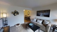 This single-floor space offers a comfortable and well-appointed layout designed for everyday living. The living room serves as the central gathering area, featuring plush carpeted flooring and crisp white walls that create a bright, clean backdrop. A contemporary gray sofa anchors the seating arrangement around a sleek black coffee table, while a decorative bowl and curated wall art add thoughtful finishing touches. A striking metallic orb wall sculpture provides an eye-catching focal point, lending the space a modern, artistic character. The living room flows seamlessly into the dining area through an open passageway, creating a connected and functional layout. The dining space features warm-toned flooring and is illuminated by overhead lighting, giving it a cozy, intimate feel. A compact dining table with matching chairs makes efficient use of the space, while a framed mirror on the wall adds depth and brightness. Overall, the space strikes a balance between practicality and style, with consistent décor choices that tie the rooms together into a cohesive and inviting home environment.