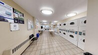 This shared laundry facility offers a clean, efficiently organized space designed to accommodate high-volume laundry needs. The room is finished with light beige walls and large-format tile flooring, creating a bright and easy-to-maintain environment. Overhead, a series of oval flush-mount light fixtures provide consistent, even illumination throughout the space.

Along one wall, a substantial bank of stacked commercial dryers lines the room, offering ample drying capacity. Opposite and toward the rear, a row of front-loading commercial washers is complemented by top-loading units, providing flexibility for various laundry loads. Instructional signage is posted near the machines to guide users on proper operation.

Central folding tables provide convenient surfaces for sorting and folding laundry. A utility sink is also present, adding further functionality. Near the entrance, a seating area with a chair, a recycling bin, and a community bulletin board with posted notices creates a practical waiting area. An additional framed artwork on the wall adds a subtle decorative touch.

The space benefits from natural light through a row of windows positioned above the washers at the rear, supplementing the artificial lighting. The layout is thoughtfully arranged to ensure smooth traffic flow, making this laundry room a functional and user-friendly shared amenity.